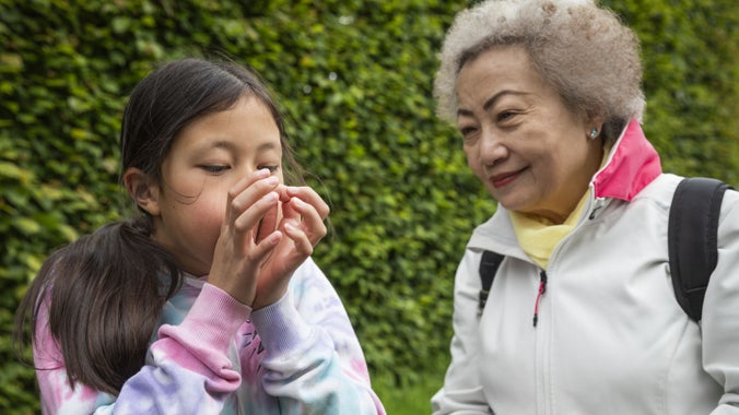 Child blows into a blade of grass in their hands while an older adult looks on, standing outdoors in front of green foliage.
